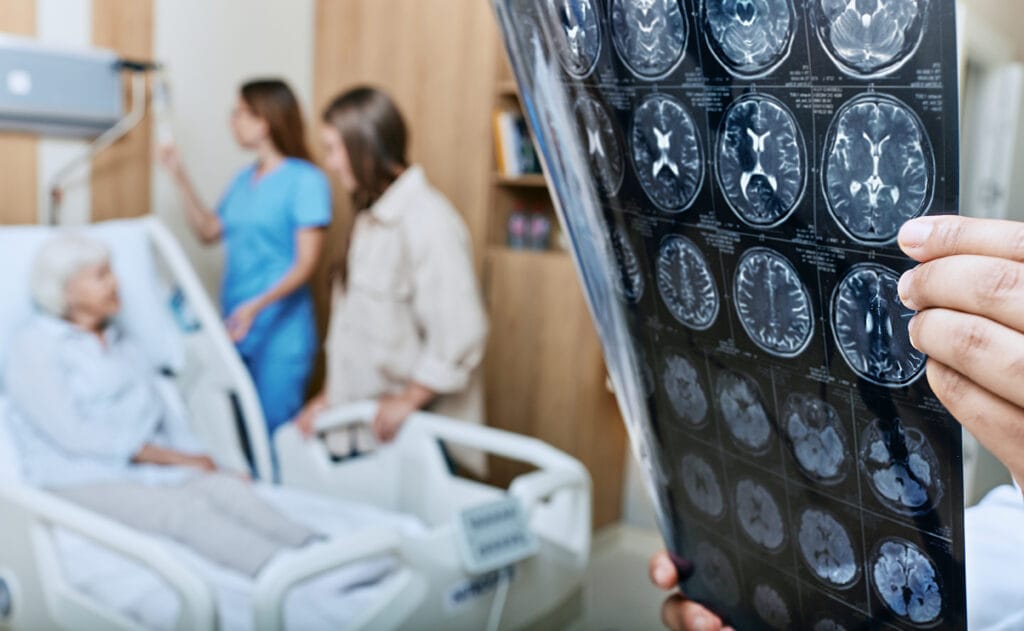 A doctor examines brain scan images while a stroke patient lies in a hospital bed, accompanied by a nurse and family member.