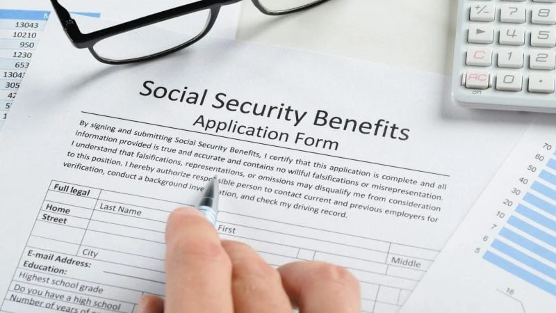 Close-up of a person filling out a Social Security benefits application form with a pen, surrounded by financial documents, charts, and a calculator.