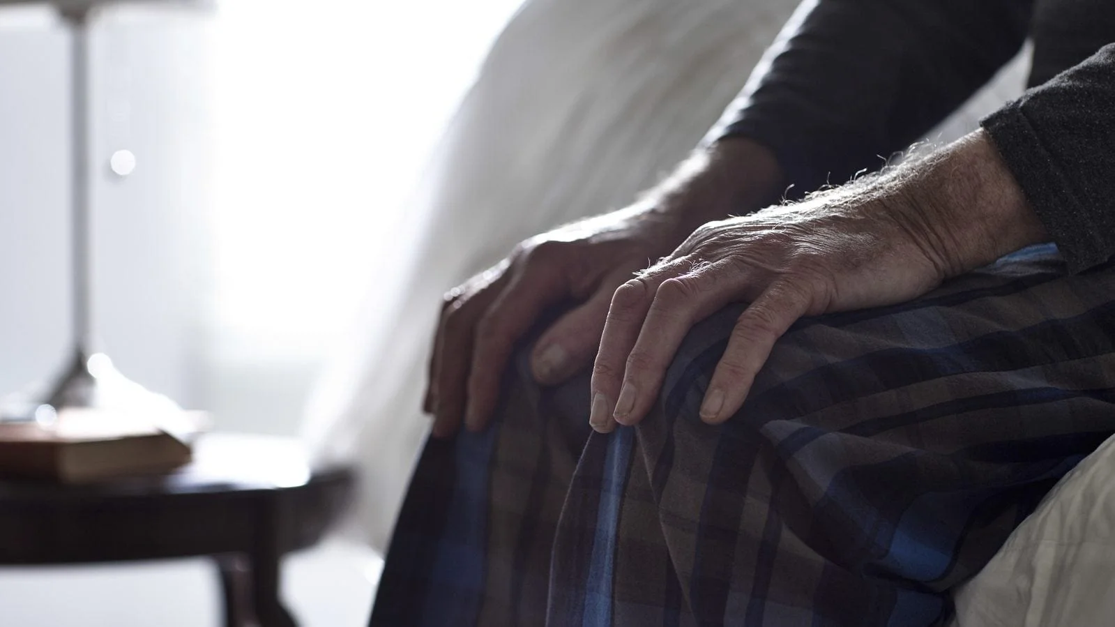 Close-up of an elderly person's hands resting on their lap while sitting on the edge of a bed.