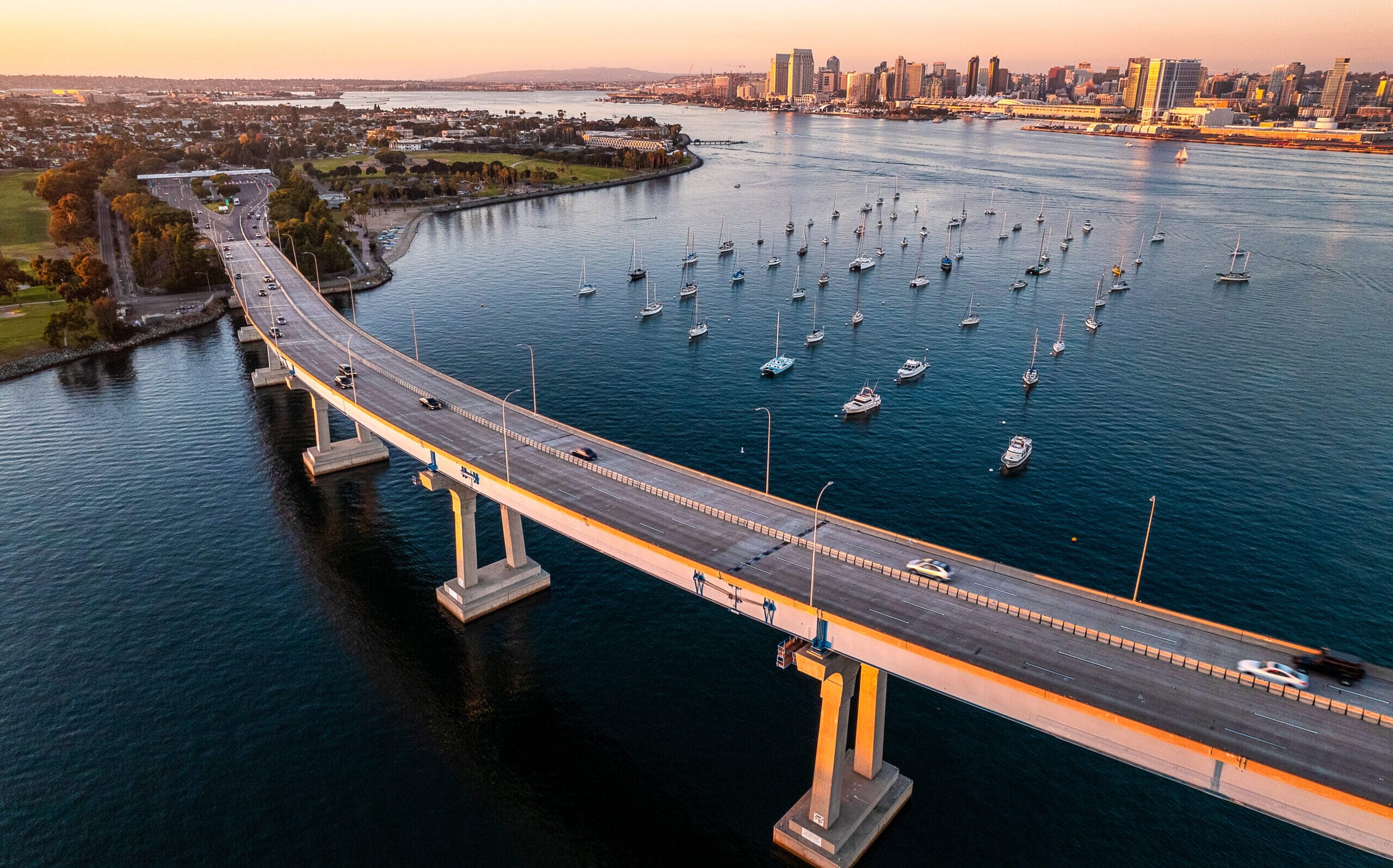 Aerial view of the San Diego–Coronado Bridge stretching over the bay with boats anchored in the water and the San Diego skyline in the background at sunset.