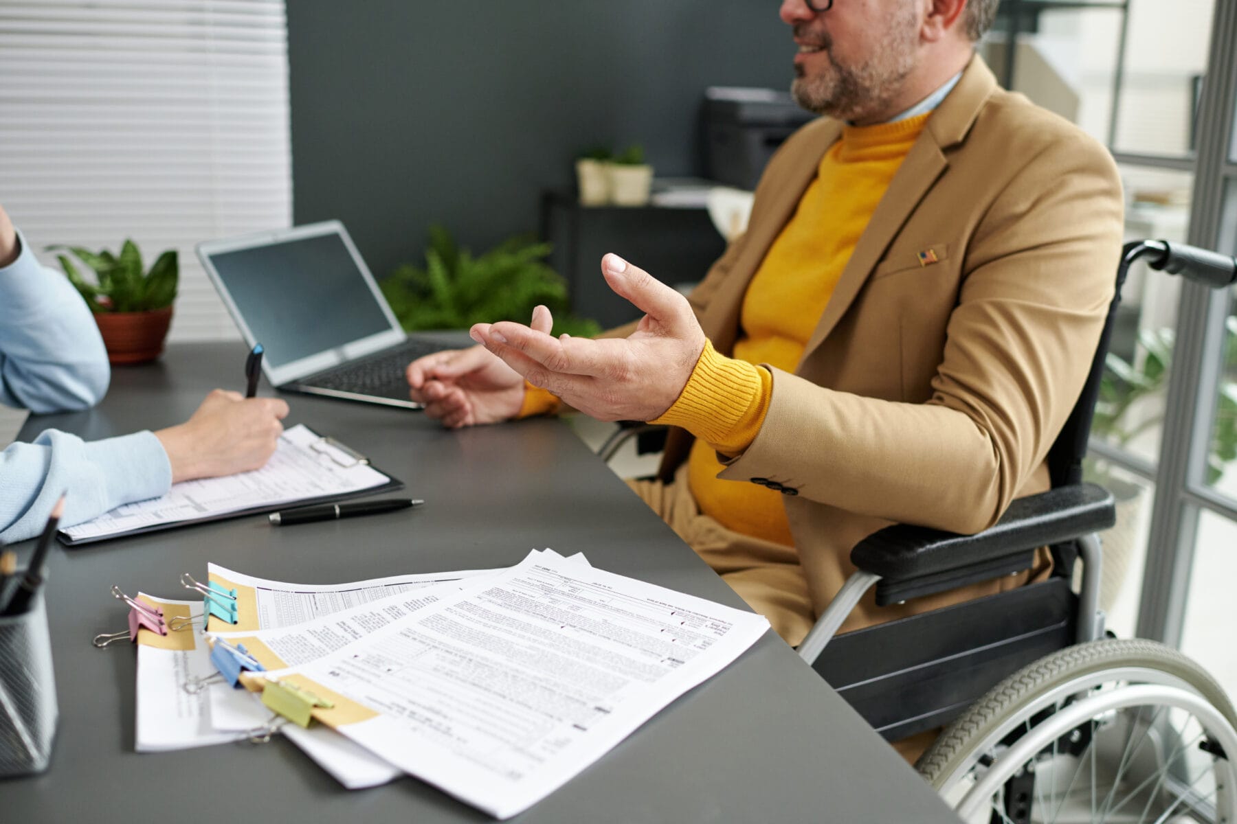 A man in a wheelchair wearing a tan jacket and yellow sweater speaks with a professional across a desk covered in paperwork.
