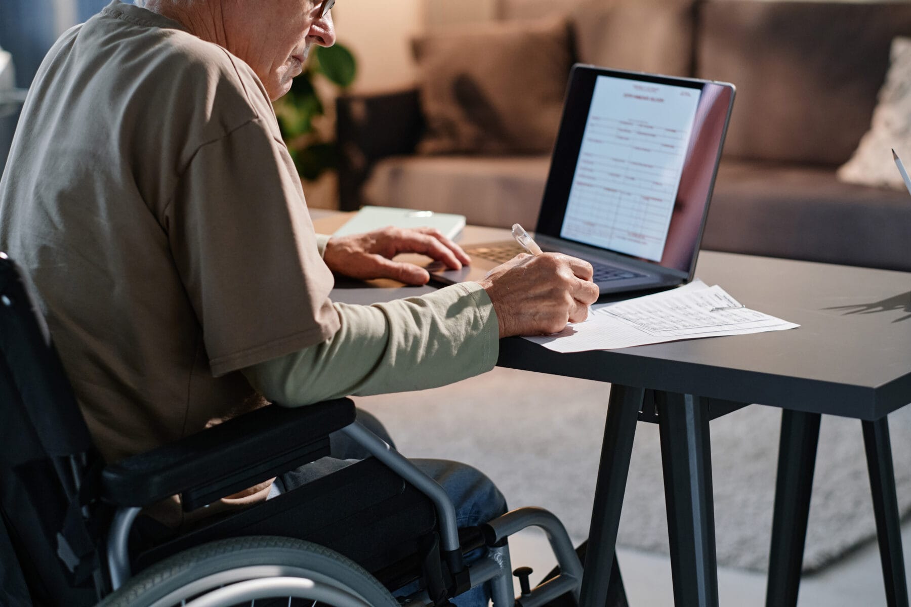 An older man in a wheelchair fills out paperwork at a desk while referencing forms on his laptop.