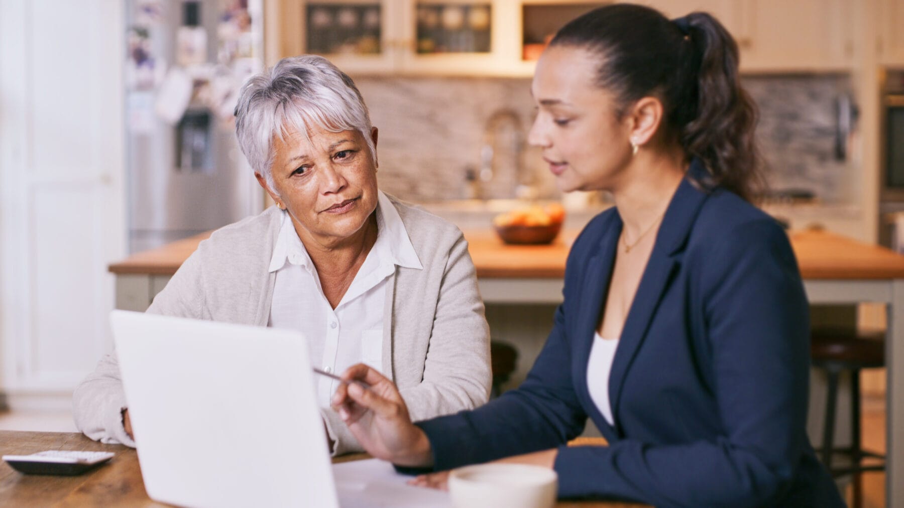 A professional woman pointing to a laptop screen with a pen, speaking with an older woman at a kitchen table.