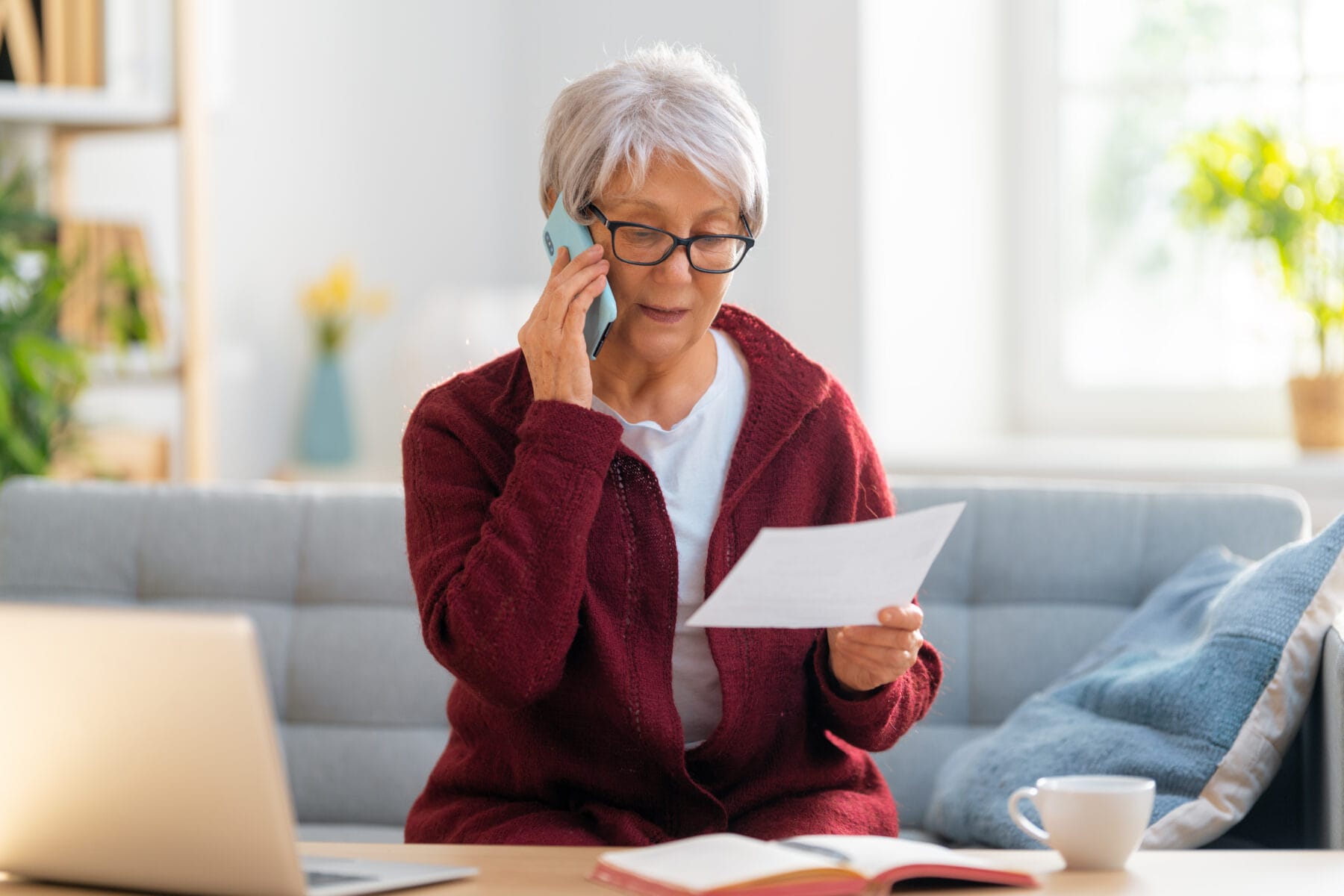 Older woman sitting on a couch talking on the phone while reviewing a document, with a laptop and coffee cup on the table in front of her.
