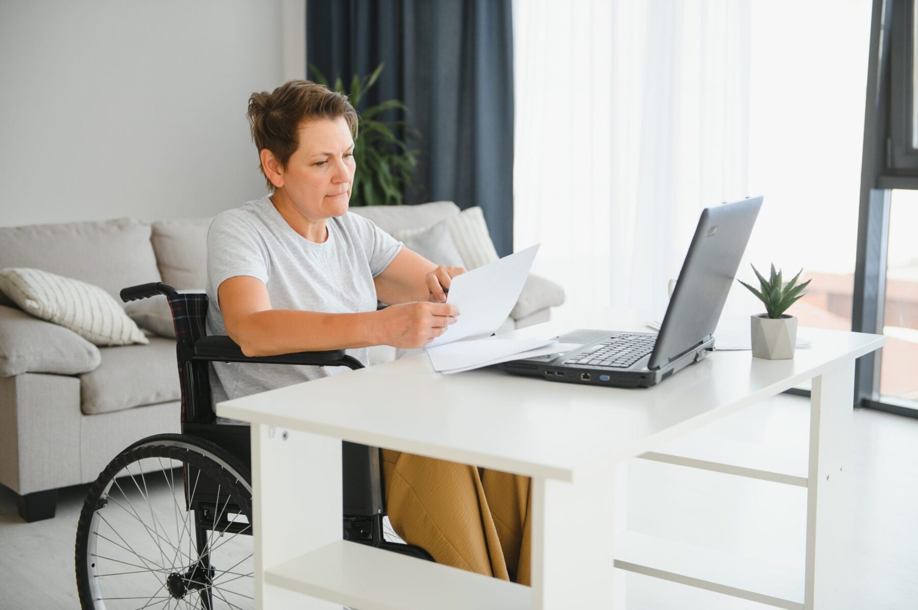 Middle-aged woman in a wheelchair reviewing SSDI paperwork at a desk while using a laptop.