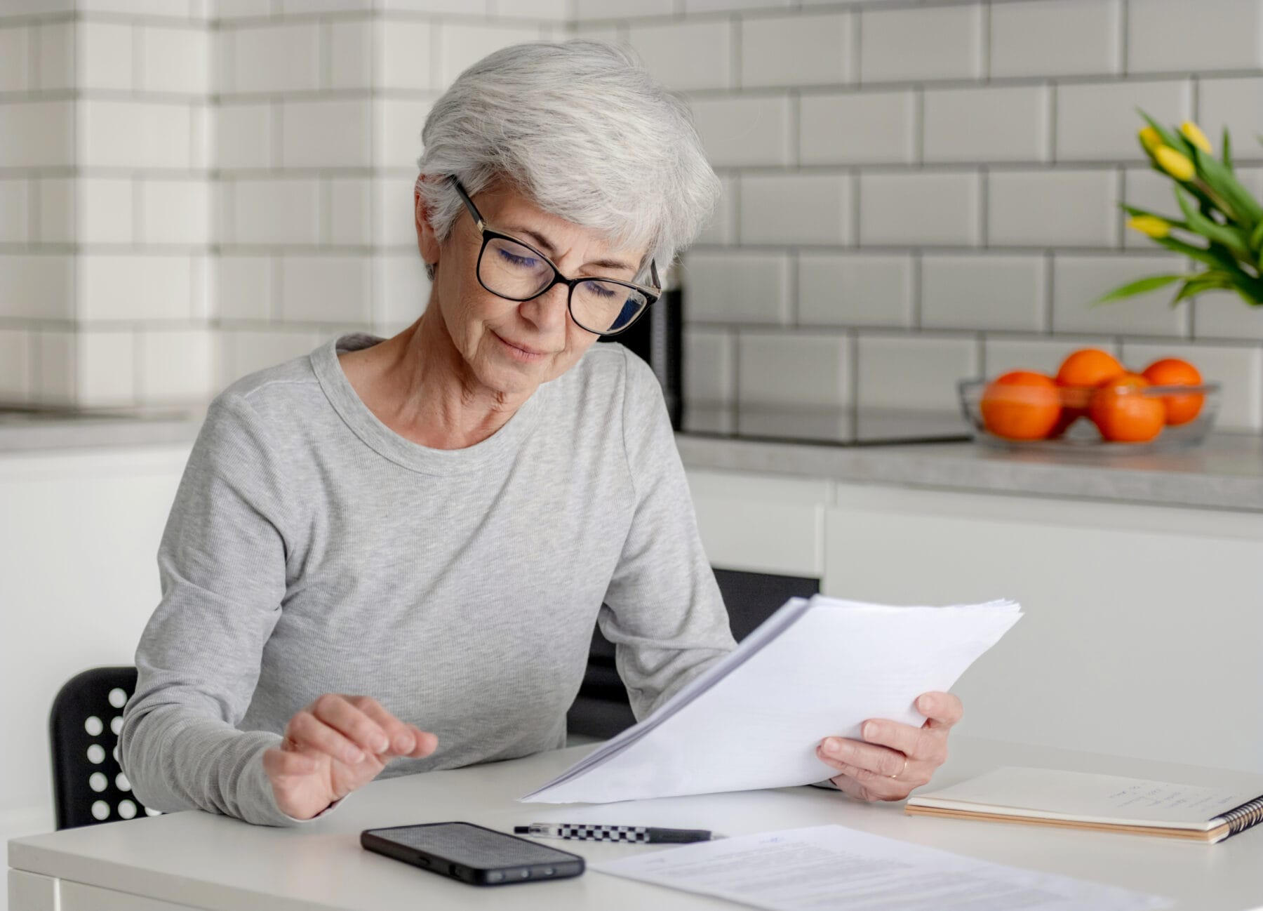 Gray-haired woman wearing glasses reviewing SSI paperwork at a kitchen table with a phone and notepad nearby.
